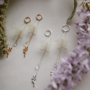 May include: Close-up of three pairs of delicate earrings. Each earring features a small, white, flower-shaped element, a chain, and a decorative charm. Two pairs have gold-colored hoops and chains, while the third pair has silver-colored hoops and chains.