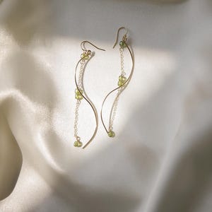 May include: A pair of gold-colored earrings with a flowing, curved design. Each earring features a delicate chain with small, light green beads. The earrings are displayed against a soft, white satin background.