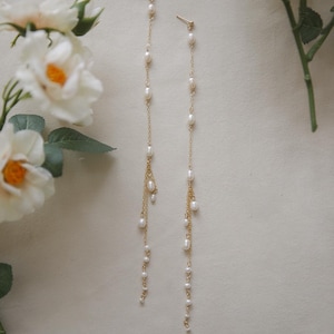 May include: A pair of elegant gold-coloured earrings. Each earring has a post and a long chain, embellished with small, lustrous pearls. The earrings hang gracefully, displaying a cascade of pearls. White flowers are in the background.