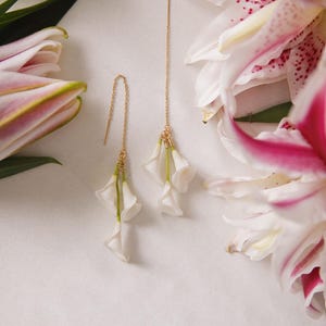 May include: Gold-toned threader earrings with white flower pendants. The earrings are displayed with white and pink lily flowers. The earrings have a delicate design, with the flower petals appearing to be made of fabric.