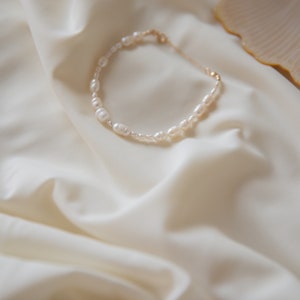 May include: A delicate pearl necklace with irregular shaped pearls on a white fabric background.