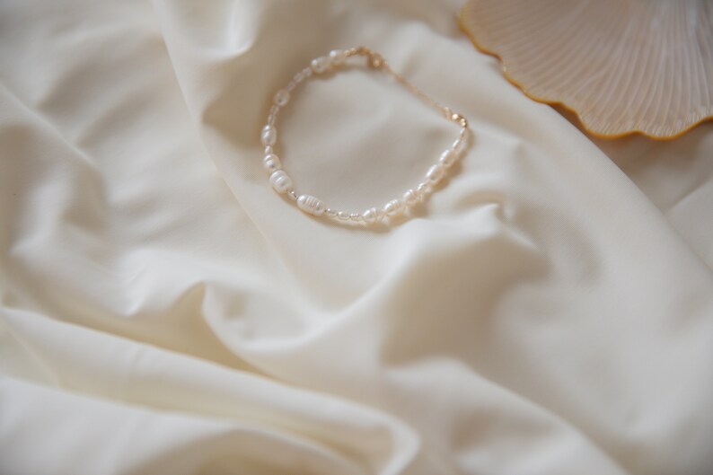 May include: A delicate pearl necklace with irregular shaped pearls on a white fabric background.