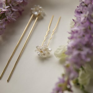 May include: Gold-coloured hair pins with pearl accents, arranged on a white surface, are surrounded by soft purple flowers. The pins include two straight pins and a wavy pin with a cluster of pearls.