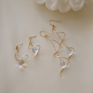 May include: Gold-toned earrings with a delicate, swirling design. Each earring features a clear, teardrop-shaped crystal pendant. One pair is a stud, the other is a long dangle style. A white flower is in the background.