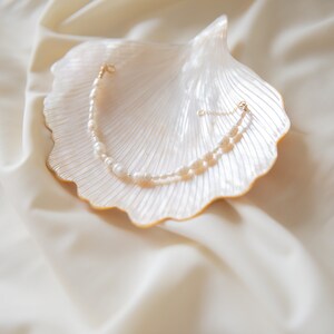 May include: A delicate pearl necklace with a gold clasp, displayed on a white seashell. The necklace is made of small, white pearls and is arranged in a simple, elegant design.