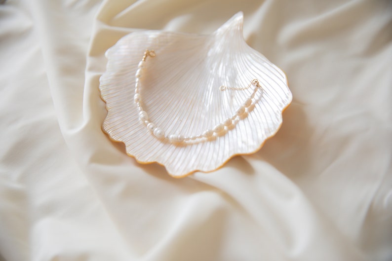 May include: A delicate pearl necklace with a gold clasp, displayed on a white seashell. The necklace is made of small, white pearls and is arranged in a simple, elegant design.