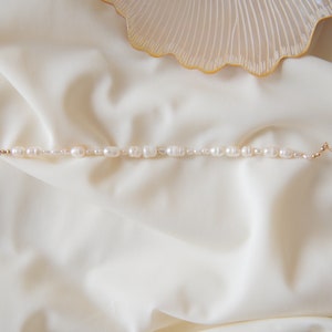 May include: A delicate pearl bracelet with a gold chain and clasp. The pearls are white and have a slightly irregular shape. The bracelet is lying on a white fabric background.