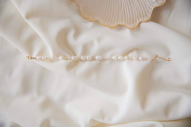 May include: A delicate pearl bracelet with a gold chain and clasp. The pearls are white and have a slightly irregular shape. The bracelet is lying on a white fabric background.