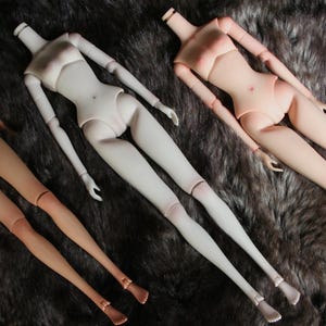 Könnte beinhalten: Drei nackte Puppen mit verschiedenen Hauttönen, die auf einer flauschigen braunen Oberfläche liegen. Die Puppen sind mit ausgestreckten Gliedmaßen posiert.