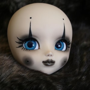 May include: A doll with blue eyes and black eyeliner, wearing a clown-like makeup design. The doll has a pale complexion and a black and white makeup design around the eyes.