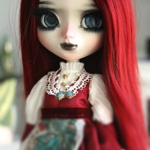May include: A doll with long, vibrant red hair and large blue eyes. The doll wears a white blouse with a red vest and a decorative collar. The doll's makeup includes dark lipstick and long eyelashes. The doll's dress has a floral pattern.