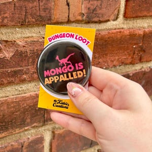 May include: A black button with the text "MONGO IS APPALLED!" in pink and orange, and a pink dinosaur illustration. The button is on a yellow card that says "DUNGEON LOOT" and "Kate's Creations".