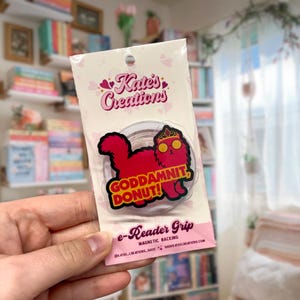 May include: A clear e-reader grip with a red cat illustration wearing a crown and glasses, with the text "GODDAMNIT, DONUT!". The grip is packaged in a pink and white card with the text "Kate's Creations".