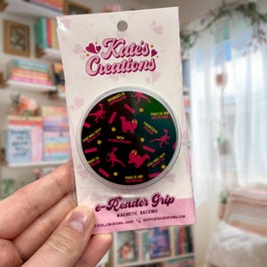 May include: A round e-reader grip with a black background and pink dinosaur illustrations. The grip has text that reads "Kate's Creations", "Mongo is Appalled", "This is an Outrage", and "Princess Posse". The grip has a magnetic backing.