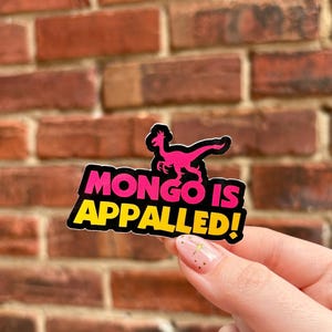 May include: A sticker with the text "MONGO IS APPALLED!" in pink and yellow, with a pink dinosaur illustration. The sticker is held in front of a red brick wall.