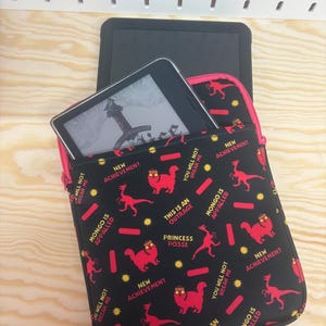 May include: A black tablet sleeve with a repeating pattern of red cartoon cats, red dinosaurs, yellow starbursts, and red rectangles. The text reads: NEW ACHIEVEMENT, YOU WILL NOT BREAK ME, MONGO IS APPALLED, THIS IS AN OUTRAGE, and PRINCESS POSSE.