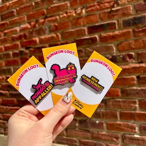 May include: Three colorful enamel pins on yellow cards with the text "DUNGEON LOOT." The pins feature phrases like "MONGO IS APPALLED!" and "GODDAMNIT, DONUT!" against a brick wall background. The pins are held by a person.