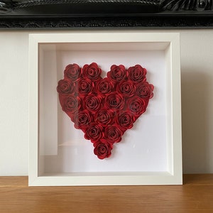 May include: A white frame with a glass front displays a red paper flower heart. The heart is made of many individual paper roses.