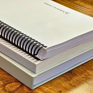 May include: A white spiral bound planner with the text "Weekly Planner" on the cover. The planner is stacked on top of a second, smaller white planner.