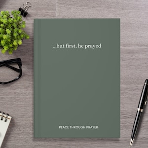 A dark sage green hardcover journal lies on a light wood desk. The front cover features minimalist white text that reads "...but first, he prayed" in the center, and "PEACE THROUGH PRAYER" near the bottom. Surrounding the journal are a black pen, black eyeglasses, a spiral notebook, a small potted green plant, and a binder clip, creating a calm and focused workspace aesthetic.
