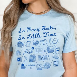 May include: A light blue t-shirt with the text "So Many Books, So Little Time" in blue lettering. The shirt is decorated with a variety of blue illustrations of books, glasses, feathers, and other book-related items.