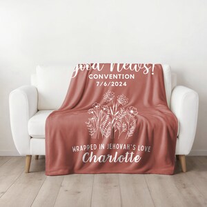 May include: A red fleece blanket with white text and a floral design. The text reads "good news! Convention 7/6/2024 Wrapped in Jehovah's love Charlotte"