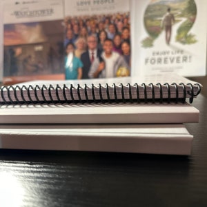 Op de afbeelding: Een close-up van een zwart spiraalgebonden notitieboek met twee witte notitieboeken die er bovenop gestapeld zijn. Het notitieboek ligt op een zwart oppervlak.