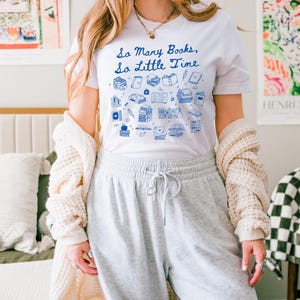 May include: A white t-shirt featuring a blue graphic of books and the text "So Many Books, So Little Time". The model is wearing the shirt with grey sweatpants and a white cardigan.