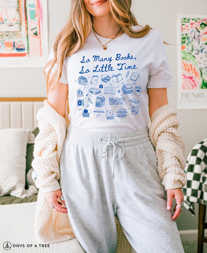 May include: A white t-shirt featuring a blue graphic of books and the text "So Many Books, So Little Time". The model is wearing the shirt with grey sweatpants and a white cardigan.