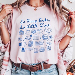 May include: A pink t-shirt with a blue graphic that says "So Many Books, So Little Time". The graphic features a variety of blue illustrations of books, glasses, and other reading-related items.