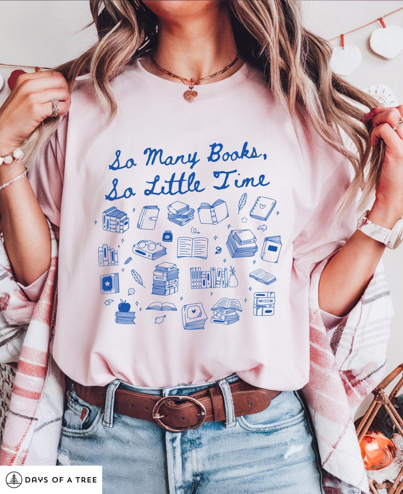 May include: A pink t-shirt with a blue graphic that says "So Many Books, So Little Time". The graphic features a variety of blue illustrations of books, glasses, and other reading-related items.