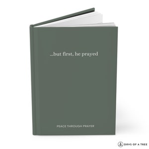 Peut inclure: Un journal &agrave; couverture rigide vert avec le texte "...but first, he prayed" et "PEACE THROUGH PRAYER" sur la couverture. Le journal est ferm&eacute; et les pages sont l&eacute;g&egrave;rement ouvertes.