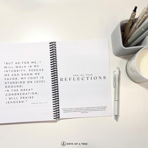 Op de afbeelding: Een wit spiraalgebonden notitieboek met de tekst "END-OF-YEAR REFLECTIONS" op de omslag. Het notitieboek is open op een pagina met een bijbelvers en gestreepte pagina's om op te schrijven. Het notitieboek ligt op een wit oppervlak met een witte pen, een kaars en een grijze pennenhouder met pennen.