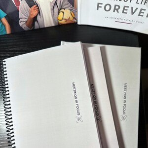 May include: White spiral-bound notebooks with the text "MEETINGS IN FOCUS" printed on the cover. The notebooks are stacked on a black surface, with a book in the background that reads "ENJOY LIFE FOREVER".
