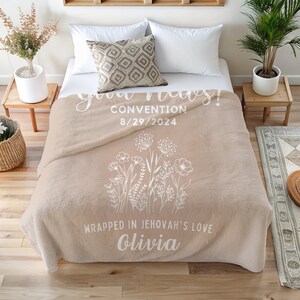 May include: A beige fleece blanket with a white floral design and the text "God bless! Convention 8/29/2024 Wrapped in Jehovah's love Olivia".