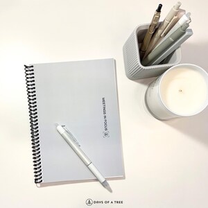 May include: A light gray spiral-bound notebook with the text "MEETINGS IN FOCUS" and a matching pen. A gray pen holder contains several pens, and a white candle sits nearby. The image is set on a white surface.