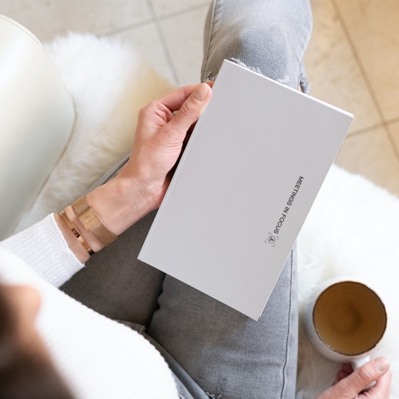 May include: A white notebook with the text "MEETINGS IN FOCUS" is held in a person's hands. The person is wearing a gold bracelet and a white sweater. A mug with a gold interior sits nearby.