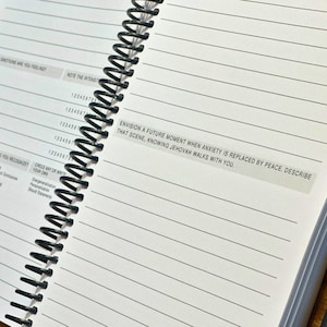 May include: A spiral bound notebook with lined pages. The notebook has a white cover with a black and white sticker that reads "Envision a future moment when anxiety is replaced by peace. Describe that scene, knowing Jehovah walks with you."