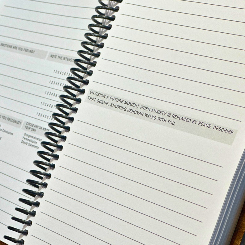 May include: A spiral bound notebook with lined pages. The notebook has a white cover with a black and white sticker that reads "Envision a future moment when anxiety is replaced by peace. Describe that scene, knowing Jehovah walks with you."