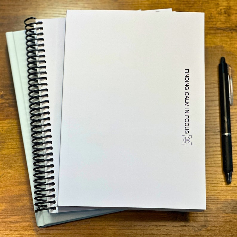 May include: A white spiral bound notebook with the text "FINDING CALM IN FOCUS" and a small square symbol with a triangle inside. The notebook is on a wooden surface with a black pen.