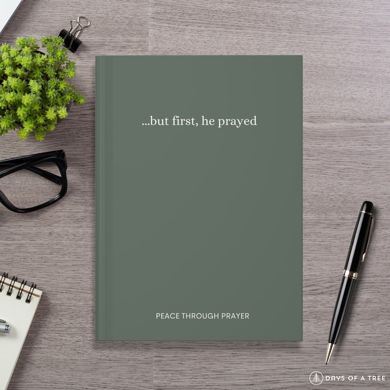 A dark sage green hardcover journal lies on a light wood desk. The front cover features minimalist white text that reads "...but first, he prayed" in the center, and "PEACE THROUGH PRAYER" near the bottom. Surrounding the journal are a black pen, black eyeglasses, a spiral notebook, a small potted green plant, and a binder clip, creating a calm and focused workspace aesthetic.
