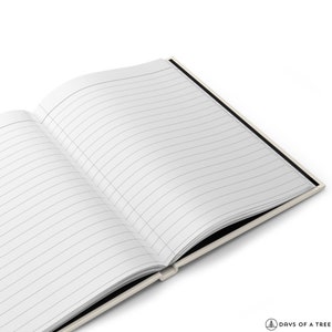 May include: An open lined notebook with a black spine and cream colored cover. The notebook is lying open on a white surface.