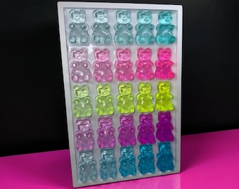 May include: A rectangular white tray containing thirty translucent gummy bear candies arranged in six rows of five. The bears are colored in gradients of blue, pink, lime green, purple, and teal, each featuring a smooth, molded bear shape.