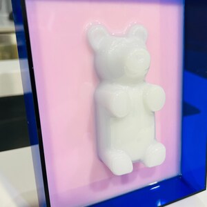 Puede incluir: Una figura tridimensional de oso de goma blanco montada sobre un fondo rosa plano, encerrada dentro de un marco rectangular azul transl&uacute;cido profundo. El oso tiene un acabado suave y brillante y est&aacute; centrado sobre el fondo rosa claro.