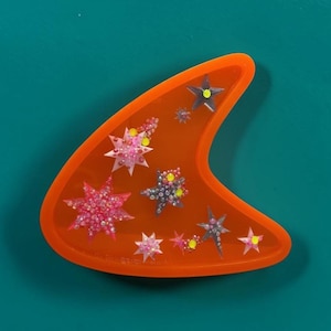Könnte beinhalten: Eine halbmondförmige orangefarbene Acrylbrosche mit eingebetteten sternförmigen Verzierungen in Rosa-, Lila- und Gelbtönen. Die Oberfläche ist glatt und durchscheinend und zeigt eine Vielzahl kleiner, mehrzackiger Sternmotive.