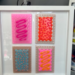 May include: A framed artwork featuring four colorful, stylized Pop-Tart designs. Each rectangular pastry has a different frosting and sprinkle pattern in shades of pink, orange, blue, and brown. The artwork is set against a white background within a white frame.