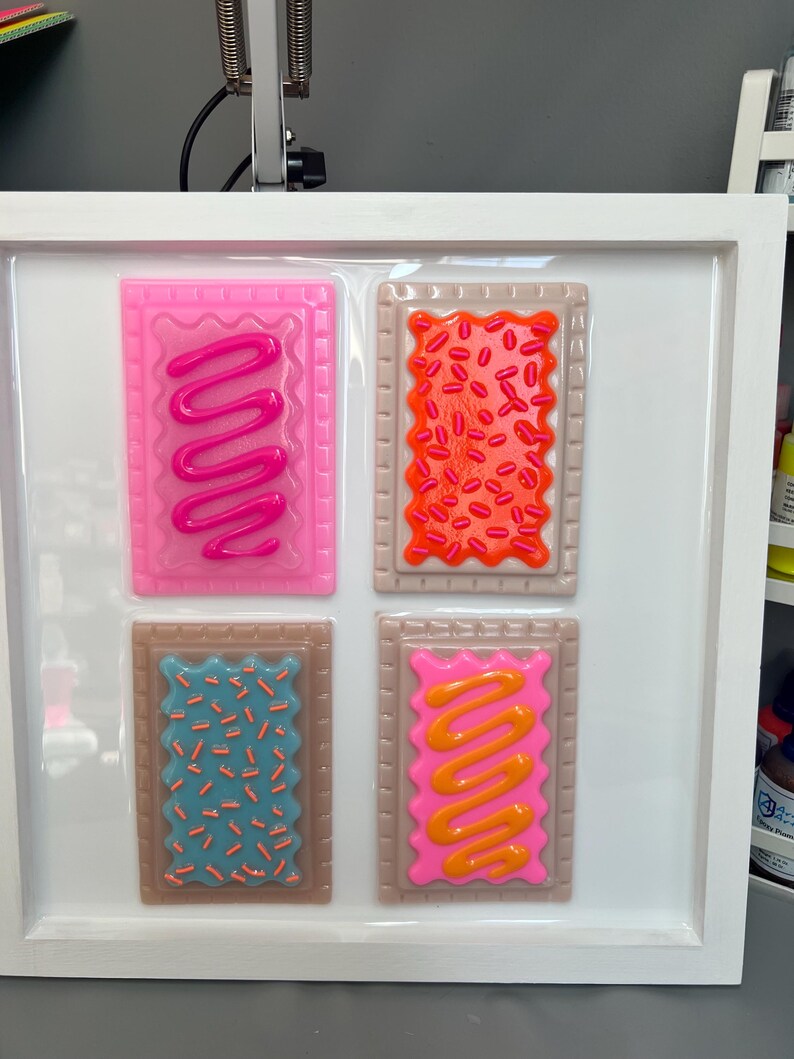 May include: A framed artwork featuring four colorful, stylized Pop-Tart designs. Each rectangular pastry has a different frosting and sprinkle pattern in shades of pink, orange, blue, and brown. The artwork is set against a white background within a white frame.