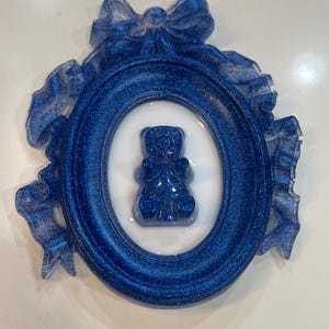Op de afbeelding: Een blauw glitterhars fotolijst met een witte ovale passe-partout en een blauwe glitterhars beer erin. De lijst is versierd met een blauwe glitterhars strik.