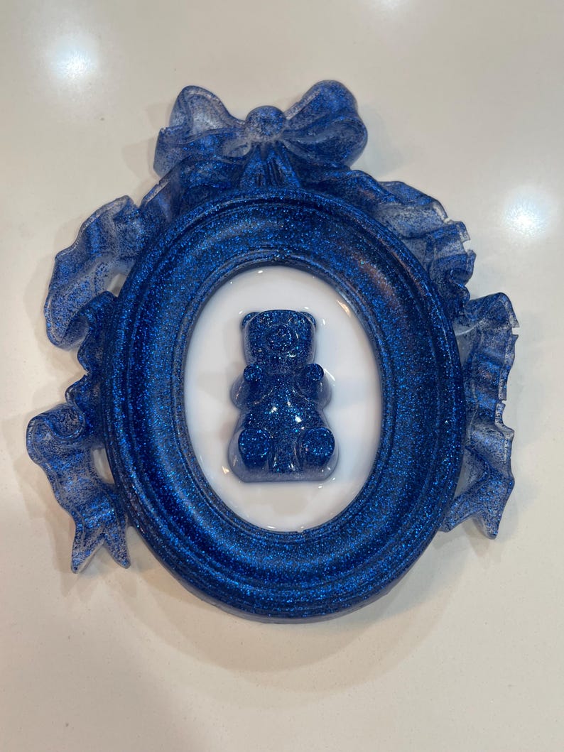 Op de afbeelding: Een blauw glitterhars fotolijst met een witte ovale passe-partout en een blauwe glitterhars beer erin. De lijst is versierd met een blauwe glitterhars strik.