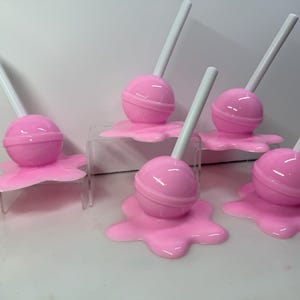 Op de afbeelding: Roze lolly's met witte stokjes, rustend op roze, gesmolten uitziende bases. De lolly's zijn gerangschikt op heldere acrylstandaards, wat een speelse en grillige esthetiek cre&euml;ert. Snoepgoed-thema tegen een witte achtergrond.
